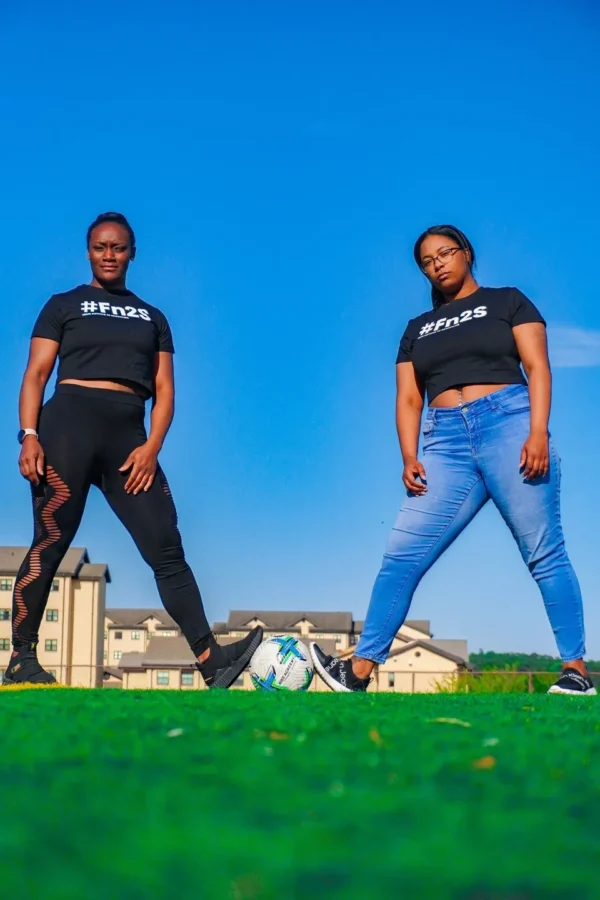 Two women standing on a soccer field in front of some buildings.